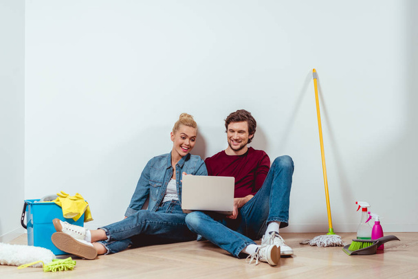 ελκυστικό κορίτσι και όμορφος άνδρας κάθεται στο πάτωμα, κοιτάζοντας οθόνη φορητού υπολογιστή και χαμογελά στο διαμέρισμα  - Φωτογραφία, εικόνα