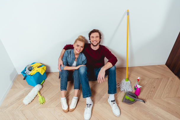 smiling couple sitting on floor near cleaning equipment and looking at camera - Фото, зображення