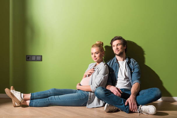 thoughtful pregnant girl sitting on floor with husband by green wall - Photo, Image