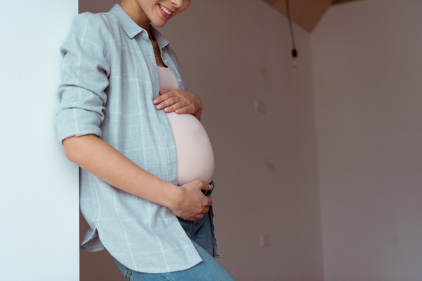 Обрезанный вид улыбающейся беременной женщины с руками на животе
 - Фото, изображение