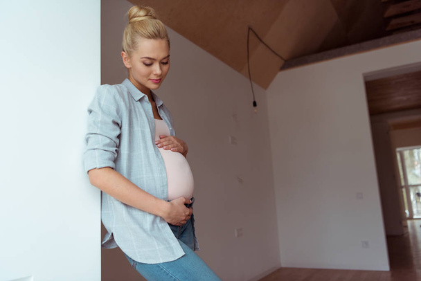 Beautiful girl holding hands on pregnant belly and standing by wall - Фото, изображение