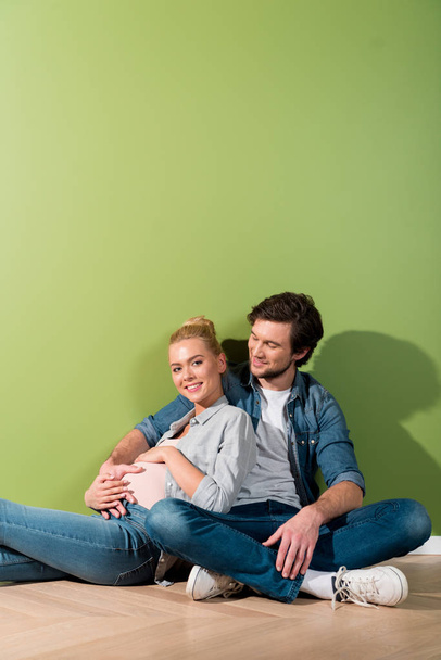 pregnant girl touching belly and sitting on floor with smiling husband by green wall - Fotoğraf, Görsel