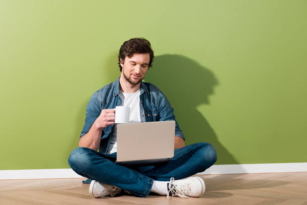 hombre guapo sosteniendo la taza de café, usando el ordenador portátil y sentado en el suelo por la pared verde
 - Foto, Imagen