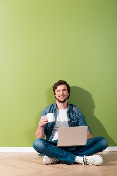 usmívající se muž držící šálek kávy a laptop a sedí na podlaze a při pohledu na fotoaparát - Fotografie, Obrázek