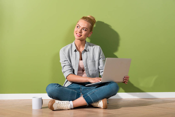 dreamy girl holding laptop and sitting on floor by green wall - Foto, imagen