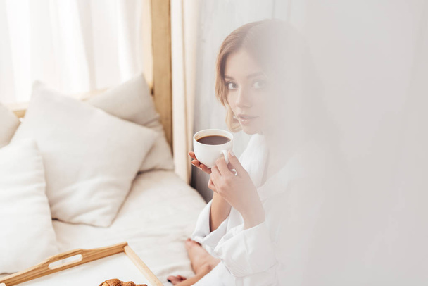 selective focus of young woman holding coffee cup and having breakfast in bed  - Zdjęcie, obraz