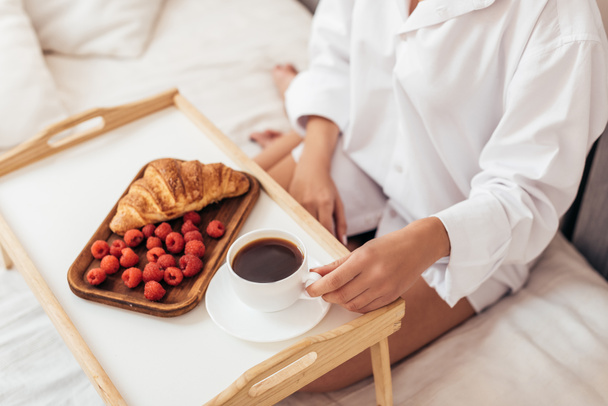 cropped image of woman having breakfast with coffee, croissant and raspberries in bed at home - Фото, изображение
