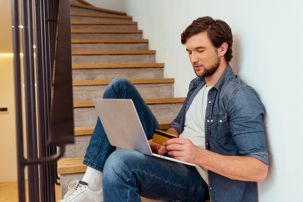 handsome man sitting on stairs, holding credit card and using laptop - Foto, afbeelding