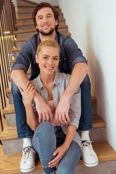 smiling man sitting on stairs and holding hands on shoulders of beautiful wife - Фото, изображение