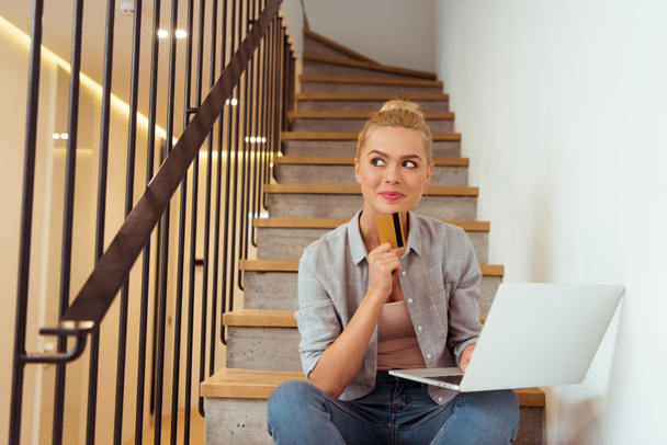 thoughtful girl sitting on stairs and holding credit card and laptop - Fotoğraf, Görsel