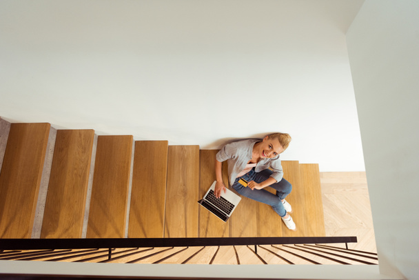 overhead view of smiling girl sitting with laptop on stairs and looking at camera - Zdjęcie, obraz