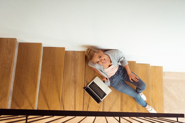zpětný pohled atraktivní dívka, sedící s notebookem na schodech a při pohledu na fotoaparát - Fotografie, Obrázek