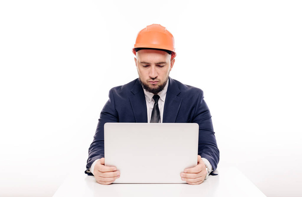 A tired constructor businessman in an orange helmet looks at the laptop screen and studies the construction project. Sits at the table and drinks coffee, nervous - Foto, Bild