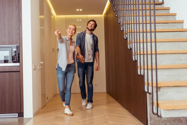 attractive girl holding hands with husband and pointing with finger near stairs at home - Φωτογραφία, εικόνα