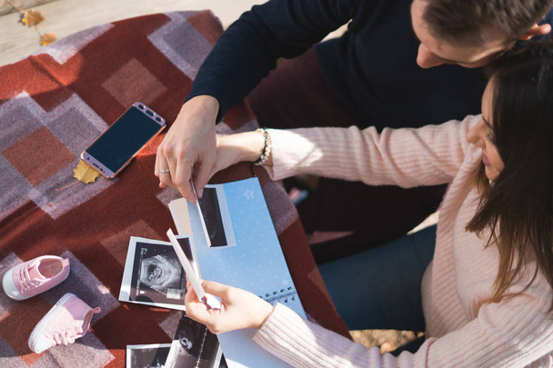 Pregnant woman and her husband makes notes in notebook and holding ultrasound image and medical documents. Pregnancy, parenthood, preparation and expectation concept.  - Fotó, kép