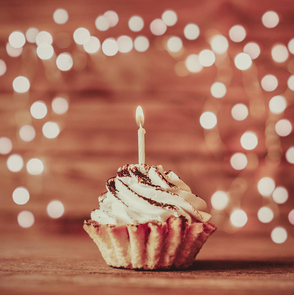 Holiday cream cupcake with burning candle on a wooden table against blurred lights. - Фото, изображение