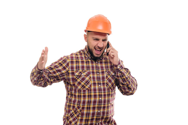 An angry and nervous worker in an orange helmet is talking loudly on the phone, shouting into the phone. Isolated white background - Foto, Imagen