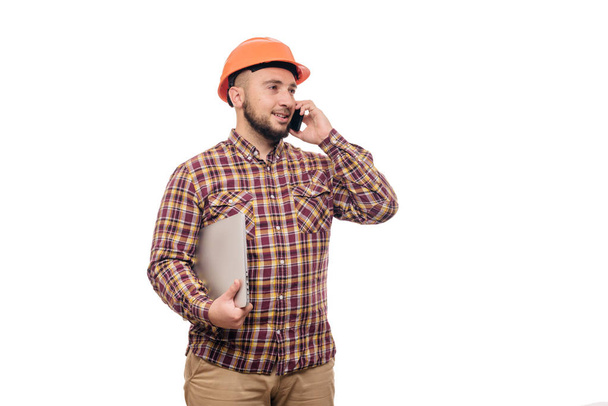 Happy Builder worker in protective construction orange helmet holding a laptop and talking on the phone, isolated on white background. Copy space for text. Time to work.  - Valokuva, kuva