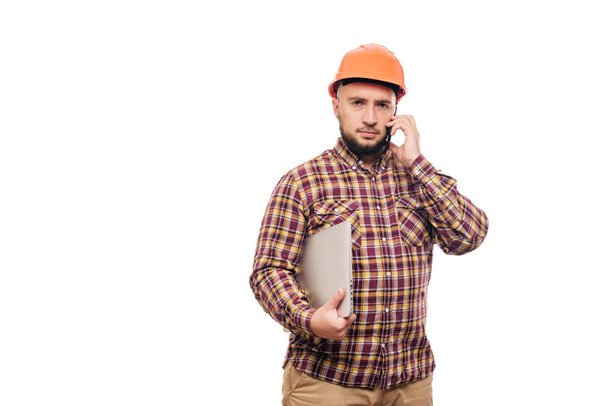 Happy Builder worker in protective construction orange helmet holding a laptop and talking on the phone, isolated on white background. Copy space for text. Time to work.  - Φωτογραφία, εικόνα