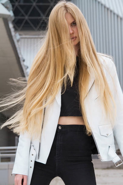 Blonde woman in the urban background. Beautiful young girl wearing a white leather jacket standing in the street. - Φωτογραφία, εικόνα