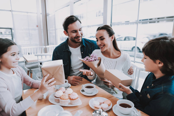Family Celebrating a Holiday in Cafe. Family is a Mother, Father, Daughter and Son. Father and Children is Giving a Flowers and Gifts to Mother. People is Happy and Smiling. Cakes and Tea on Table. - Foto, Imagen