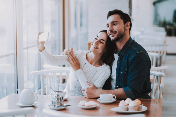 Couple Resting in Cafe. Couple is Beautiful Young Man and Woman. Couple is Doing Selfie on Mobile Phone. Persons is Sitting at Table. People is Happy and Smiling. Cakes and Tea on Table. Daytime. - 写真・画像
