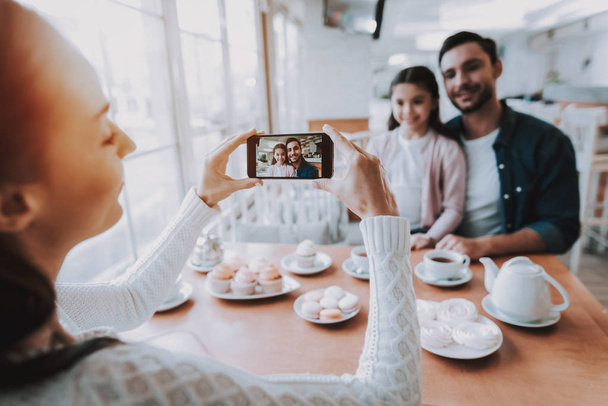 Family Resting in Cafe. Family is a Mother, Father and Daughter. Mother Photographs Father with Daughter on Mobile Phone. Persons is Sitting at Table. People is Happy and Smiling. Daytime. - 写真・画像