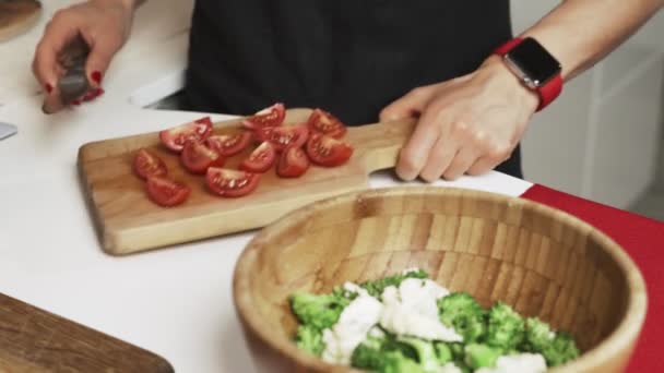 Keuken in het appartement. jonge vrouw snijdt de tomaten op een houten bord met mes, groenten op de eettafel. Cherry tomaten. - Video