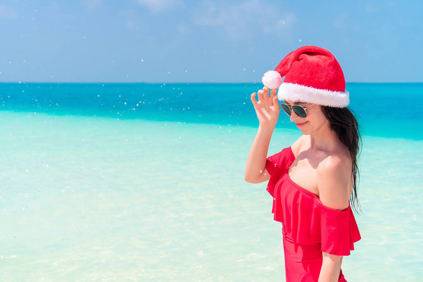 Young happy woman in Santa hat and in red swimsuit on white beach on Christmas holidays - Photo, Image