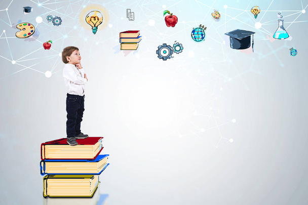 Seitenansicht des entzückenden kleinen Jungen in weißem Hemd und Jeans, der auf einem Bücherstapel neben der weißen Wand mit bunten Bildungsskizzen steht. Attrappe - Foto, Bild