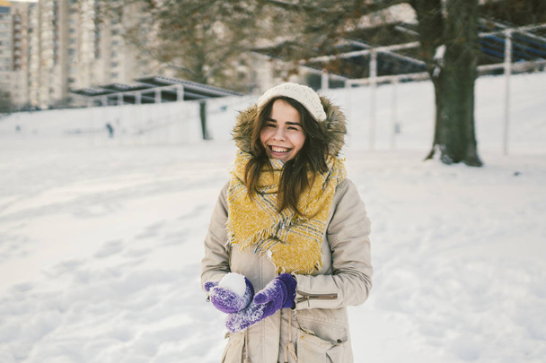 Πορτρέτο του μια όμορφη νεαρή γυναίκα Καυκάσιος σε ένα πλεκτό καπέλο και κασκόλ που στέκεται πάνω σε ένα χειμώνα φόντο με χιόνι χαμόγελο και η ευτυχία μωβ γάντια sculpt μια χιονόμπαλα. - Φωτογραφία, εικόνα