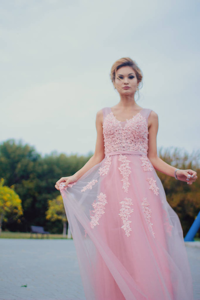Young beautiful woman in long pink evening dress walking path in park. Fashion style portrait of gorgeous beautiful girl outdoors near the Ferris wheel - Φωτογραφία, εικόνα