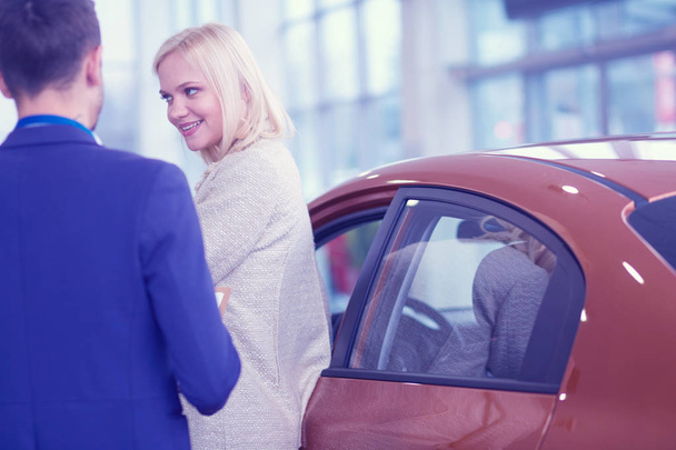 Dealer with woman stands near a new car in the showroom - Фото, изображение
