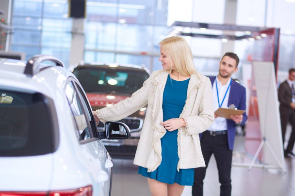Dealer with woman stands near a new car in the showroom - Zdjęcie, obraz