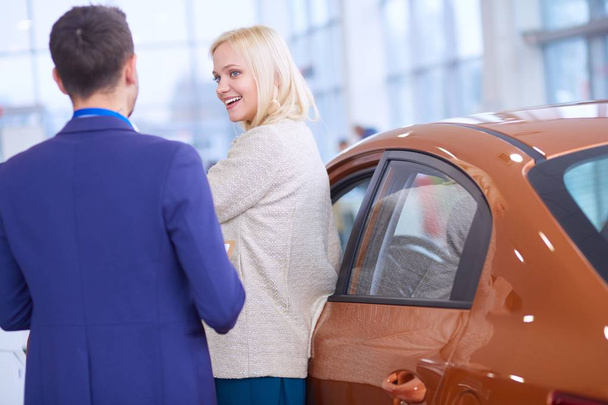 Dealer with woman stands near a new car in the showroom - Fotó, kép