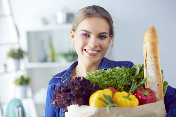 Fiatal nő gazdaság élelmiszer bevásárló táska zöldségekkel.Álló a konyhában - Fotó, kép