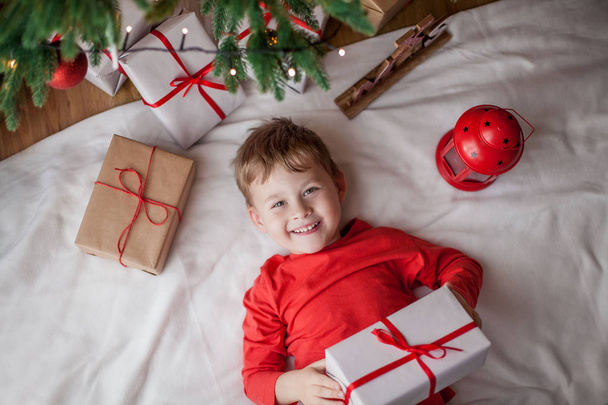 Pieni iloinen söpö poika makaa lähellä joulukuusi ja pitää lahjapakkauksessa. Lapsi. Joulutunnelmaa. Juhlapyhät. Joulu. Uusi vuosi
. - Valokuva, kuva