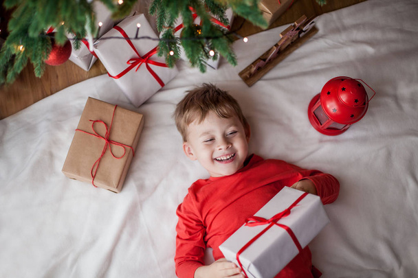 Pieni iloinen söpö poika makaa lähellä joulukuusi ja pitää lahjapakkauksessa. Lapsi. Joulutunnelmaa. Juhlapyhät. Joulu. Uusi vuosi
. - Valokuva, kuva