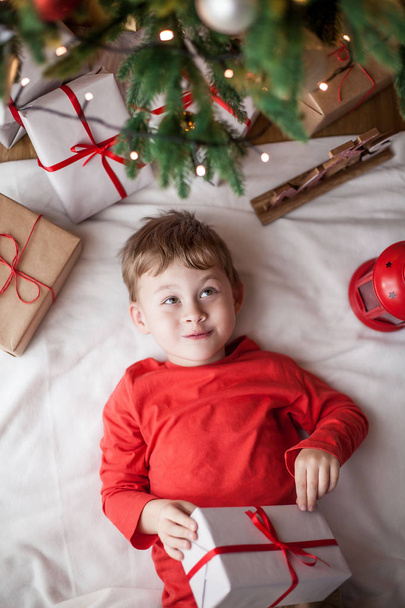 Ευτυχισμένος χαριτωμένο αγοράκι βρίσκεται κοντά το χριστουγεννιάτικο δέντρο και κρατά ένα κουτί δώρου. Το παιδί. Χριστουγεννιάτικη διάθεση. Διακοπές. Χριστούγεννα. Το νέο έτος. - Φωτογραφία, εικόνα