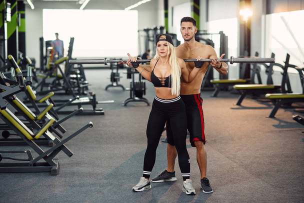 Beautiful girl doing squats under the supervision of the coach. Young couple is working out at gym. Attractive woman and handsome muscular man training together in modern gym. - Foto, imagen