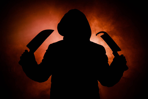 A dangerous hooded man standing in the dark and holding big knives. Face can not be seen. Committing a crime concept - Zdjęcie, obraz