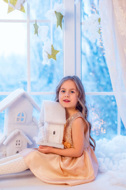 Χαριτωμένο μικρό κορίτσι με σγουρά μαλλιά στο παράθυρο εν όψει των Χριστουγέννων και της Πρωτοχρονιάς μαγεία. Αστεία συναισθήματα σε μέρα του χειμώνα. - Φωτογραφία, εικόνα