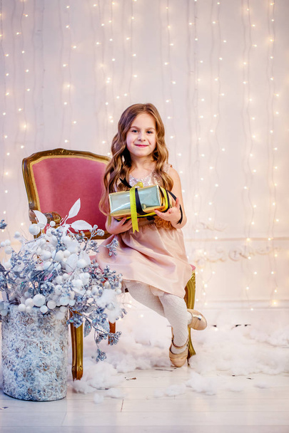 Cute little girl with curly hair with a gift on Christmas Eve, before the New Year. Cheerful emotions in anticipation of a surprise. - Fotografie, Obrázek