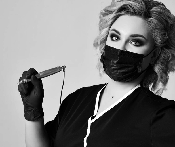 Woman beautician cosmetologist hold tattoo machine gun in black mask on gray background - Fotoğraf, Görsel