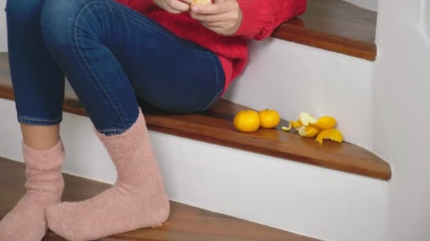 Beautiful girl in red sweater and jeans, sitting on the stairs, cleans tangerines and eats them. Christmas mood concept - Záběry, video