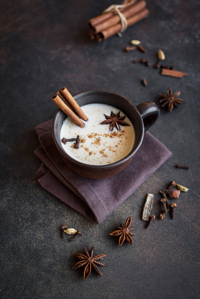 Traditional indian masala chai tea in ceramic cup with ingredients. Spicy black tea with milk (Masala) on rustic dark background. - Photo, image