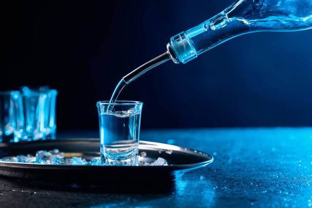 Vodka derramado em um copo iluminado com luz de fundo azul. Copiar espaço.Vodka gelada com pedaços de gelo
. - Foto, Imagem