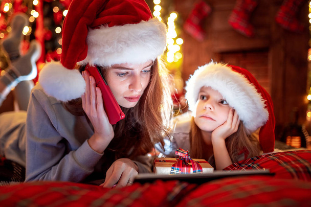 Família mãe e filha em chapéus e pijamas escolhendo presentes no tablet digital e mãe chama Papai Noel por telefone móvel
 - Foto, Imagem