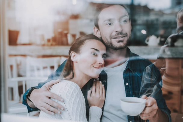 Couple Resting in Cafe. Couple is Beautiful Young Man and Woman. Couple is Looking Out from Window and Hugging. Man is Drinking a Tea. Persons is Sitting at Table. People is Happy and Smiling. - 写真・画像