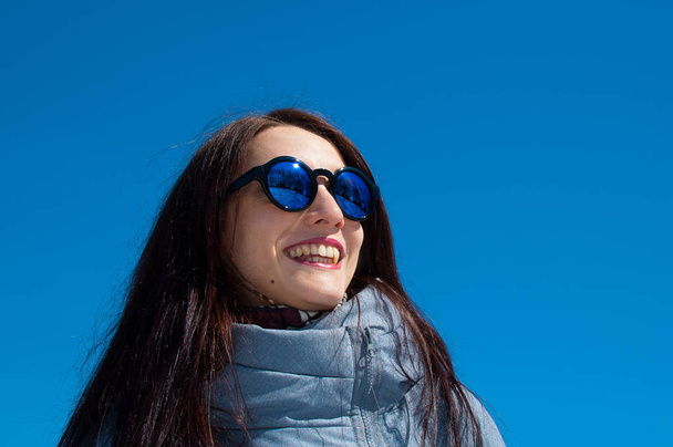 Outdoors lifestyle close up portrait of beautiful girl walking in the snowy winter park. Smiling and enjoying wintertime. Wearing stylish mirrored sunglasses, blue trench coat - Foto, immagini